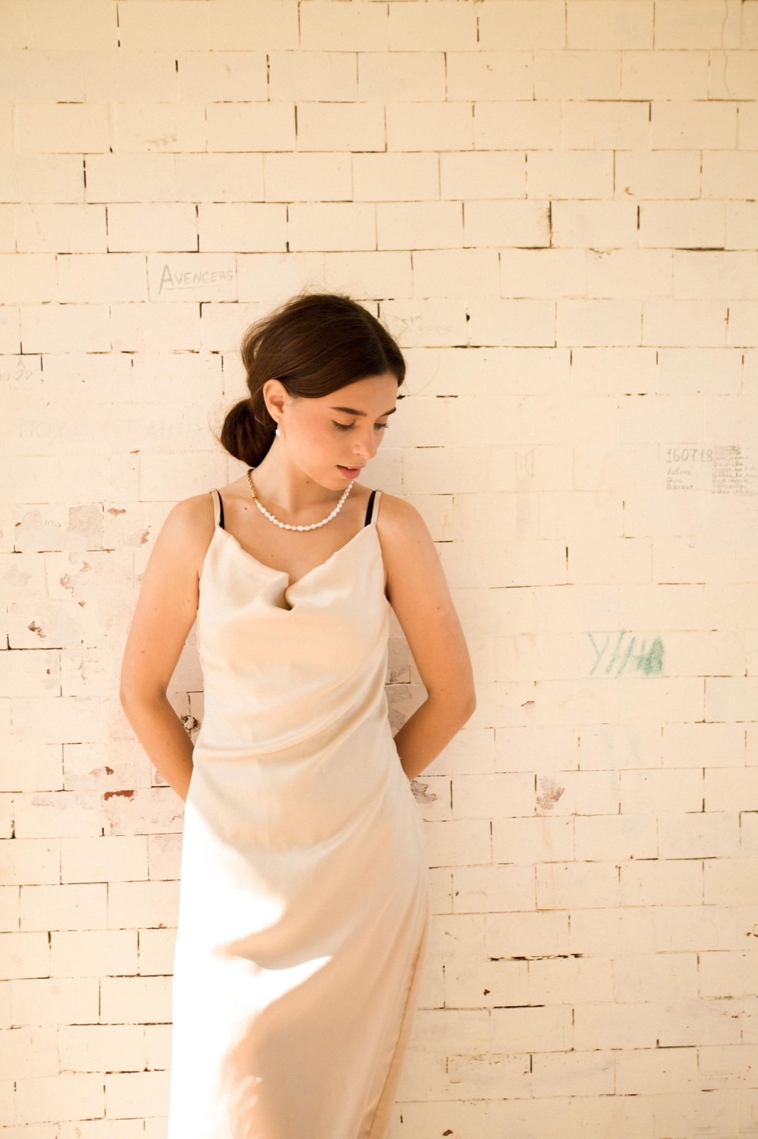 A woman in a cream dress beside a sunlit textured wall.